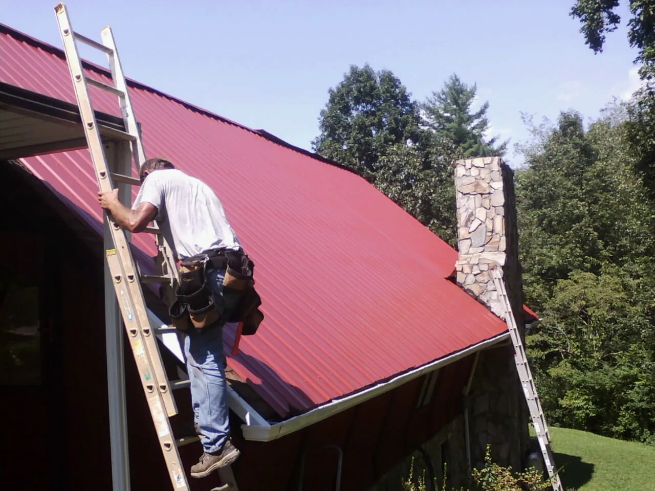 Roofer on a ladder beside a red metal roof and stone chimney — Soffit & Fascia Repair by Roofing Brothers in Bear Lake